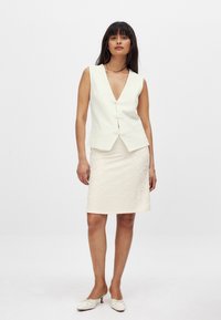 Gilet blanc sans manches avec un col en V et une fermeture décorative à boutons, associé à une jupe crayon crème texturée, porté avec des chaussures à talons blancs.