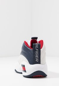 Sneaker o masywnej konstrukcji w kolorach białym, granatowym i czerwonym. Gładkie i teksturowane materiały, wyraźne logo na pięcie, pętla do zakupu.