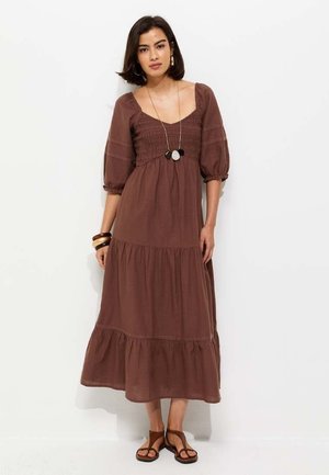 Donna che indossa un vestito lungo marrone a balze, con maniche a sbuffo e scollo quadrato, accessoriato con braccialetti, collana e sandali.