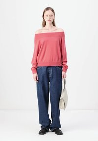 Pull rose décolleté, jeans en denim bleu ample, chaussures noires, tenant un sac gris clair ; tissu lisse, coupe décontractée, style casual.