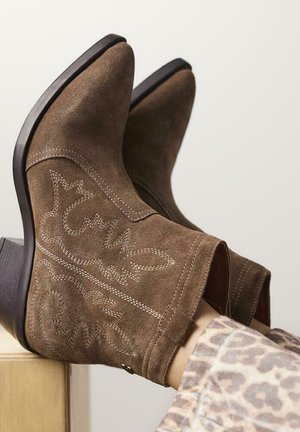 Botas de tobillo de ante marrón con punta afilada y tacón cuadrado. Presentan costuras decorativas en hilo claro y acentos de diseño recortados.