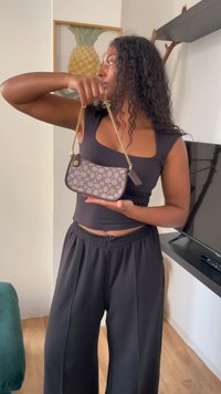Vrouw met krullend haar in een zwarte top en broek, die een kleine handtas met een gouden schakelband vasthoudt in een woonkamer.