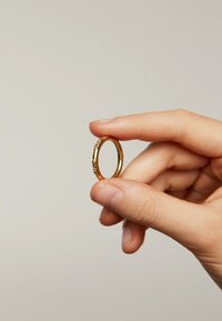 Anillo de oro sostenido entre los dedos, con una superficie lisa y pequeños acentos decorativos a lo largo del borde. Forma redonda, acabado pulido.