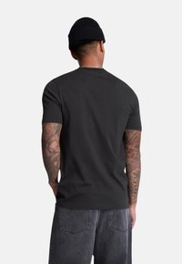 Tricou negru din bumbac, cu mâneci scurte, tăietură slim, cu guler rotund. Purtați cu pantaloni din denim gri, având tatuaje vizibile pe brațe.