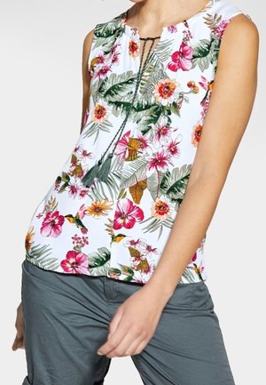 Mouwloze witte blouse met kleurrijke tropische bloemen- en vogelprint, strik met franjes bij de hals, gedragen met grijze broek.