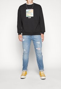 Hombre con sudadera negra de Levi's con estampado de montaña, jeans rotos de color azul claro y zapatillas altas amarillas.