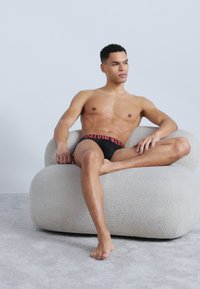 Άνδρας χωρίς πουκάμισο που φοράει μαύρο εσώρουχο Calvin Klein, καθισμένος σταυροπόδι σε μια στρογγυλεμένη, υφασμένη λευκή πολυθρόνα πάνω σε γκρι χαλί.