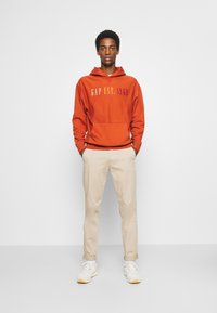 Sudadera naranja con capucha con el texto "GAP EST. 1969" impreso en colores degradados, combinada con pantalones chinos beige y zapatillas blancas. Fondo liso.