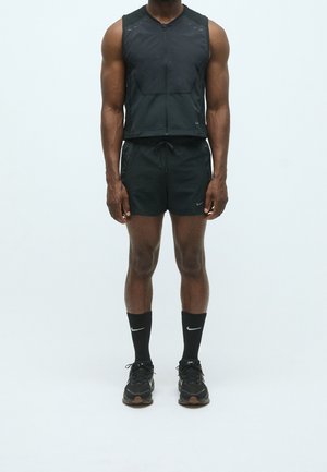 Mann trägt schwarze ärmellose Zip-Weste, schwarze Shorts mit Kordelzug, schwarze Nike-Socken und schwarze Laufschuhe, steht vor schlichtem Hintergrund.