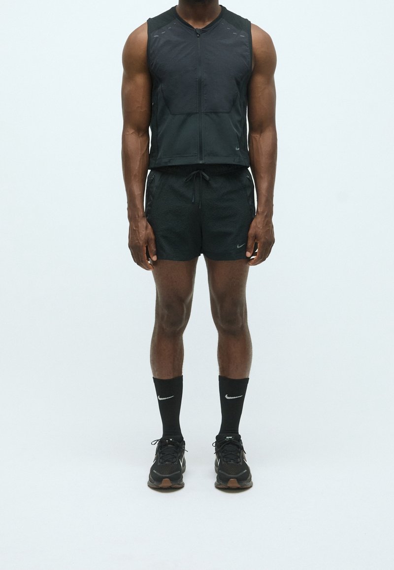 Homme portant un gilet noir sans manches zippé, un short noir à cordon, des chaussettes Nike noires et des chaussures de course noires, debout devant un fond uni.