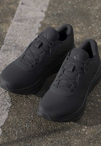 Paire de baskets de sport noires avec lacets sur une surface d'asphalte rugueuse avec une ligne blanche fissurée à proximité.
