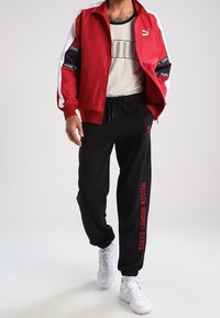 Chaqueta de satén roja con acentos blancos y azul marino, camiseta de malla negra, pantalones negros con texto rojo y zapatillas blancas. Tejido texturizado y estilo deportivo.