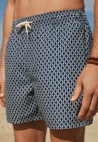 Pantaloni da bagno blu navy con un motivo geometrico bianco, vita elastica e cordino. Texture liscia, lunghezza fino a metà coscia.