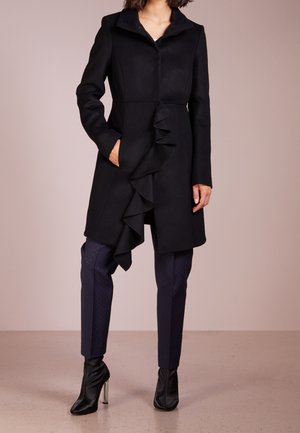 Kobieta ubrana w czarny płaszcz z falbanami z przodu, granatowe dopasowane spodnie z subtelnymi kropkami oraz czarne wysokie botki na obcasie, stojąca na jednolitym tle.