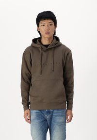 Brun bomulds hoodie med kængurulomme, snoretræk og frontlogo. Kombineret med lysvaskede denim jeans. Modellen står foran en hvid baggrund.