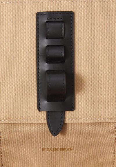 Beige fabric bag with black leather strap accents and closures. Features visible stitching and a brand text embossed at the bottom.