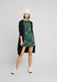 Svart cardigan over en grønn blomstret kjole, kombinert med hvite joggesko. Cardiganen har lange ermer og åpen front; kjolen har en tettsittende overdel.