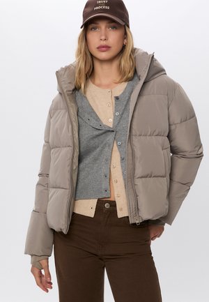 Beige puffjacka med huva, mjukt material, dragkedja, lagd över en grå cardigan och en beige t-shirt, kombinerad med bruna byxor.