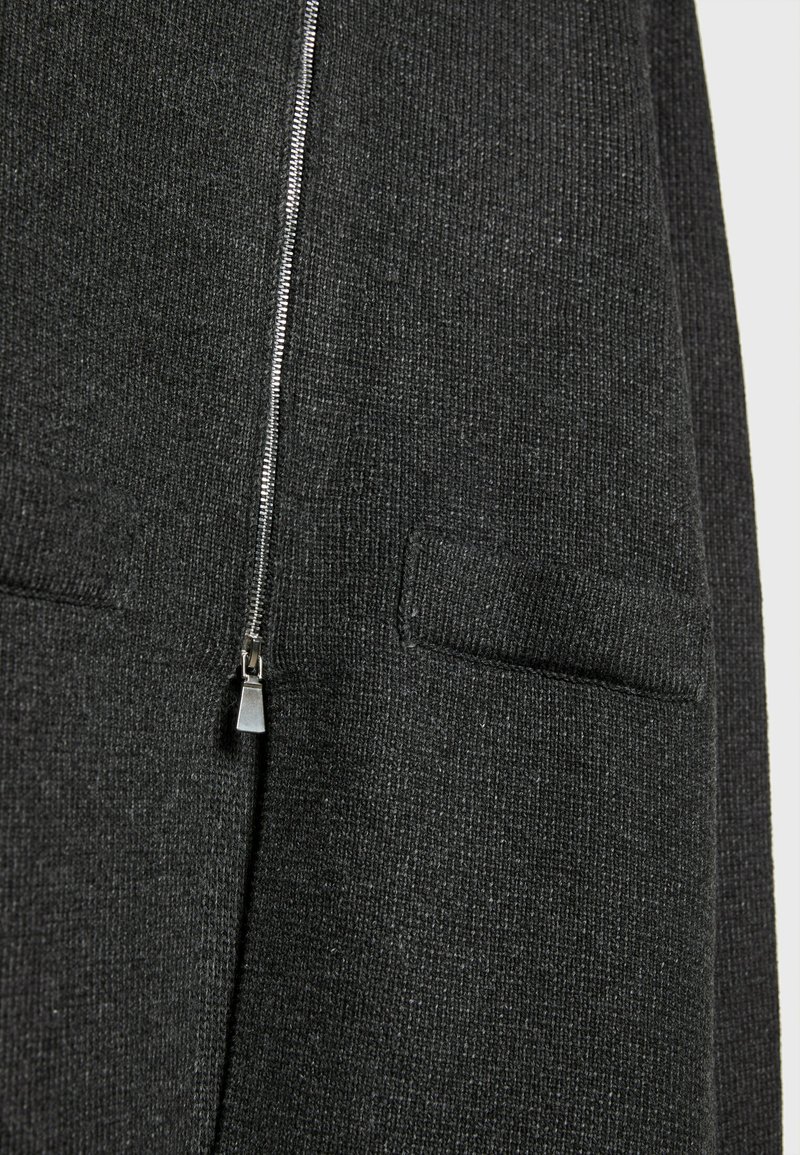 Cardigan en tricot gris foncé avec une fermeture éclair à l'avant, doté de deux poches latérales. Le tissu semble texturé. La tirette de la fermeture éclair est métallique.