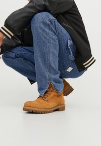 Botas de trabajo de cuero marrón con acabado texturizado y suelas de goma, combinadas con un jean de mezclilla azul claro. La persona está agachada.