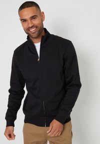 Zwarte zip-up sweatshirt met hoge kraag, lange mouwen, geribbelde manchetten en tailleband. Zachte textuur, eenvoudig ontwerp, gedragen over een witte t-shirt.