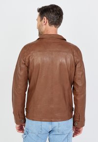 Eine braune Lederjacke mit klassischem Kragen, gesteppten Details auf den Schultern und Druckknöpfen an den Ärmelbündchen und der Taille, getragen mit hellblauen Jeans.