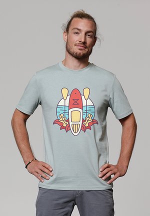 Mann mit zurückgebundenem Haar, der ein hellblaues T-Shirt mit einem bunten Design aus Kajak, Paddeln, Wasser und Korallen trägt. Hände in die Hüften gestützt.