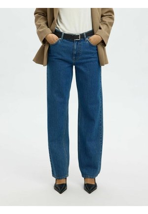 Osoba nosiaca modré džínsy s rovnými nohavicami, čierny opasok, béžové sako, biely top a čierne špicaté topánky, ruky vreckách.