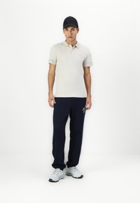 Lys beige polo skjorte med kortærmer og logo detaljer, parret med marineblå sweatpants og grå sportsko. En sort kasket fuldender outfittet.