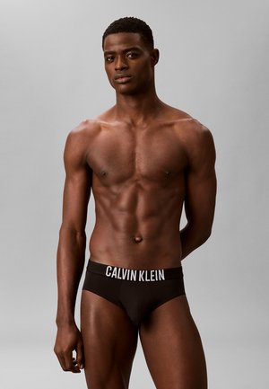 Uomo muscoloso in piedi davanti a uno sfondo grigio, indossa slip neri Calvin Klein, guarda avanti con un'espressione neutra.