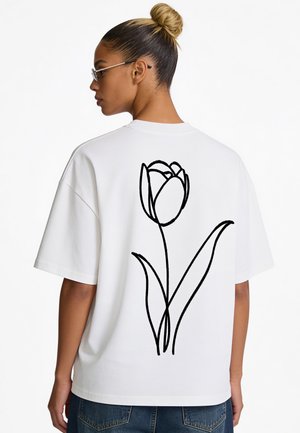 Frau mit blondem Haar zu einem Dutt gebunden, trägt eine Brille und ein weißes T-Shirt mit einer großen schwarzen Linienzeichnung einer Tulpe auf dem Rücken.
