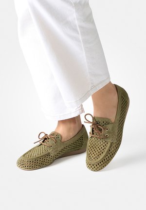 Füße, die olivgrüne perforierte Wildleder-Loafer mit braunen Schnürsenkeln tragen, kombiniert mit weißen, gekürzten Hosen vor einem schlichten Hintergrund.