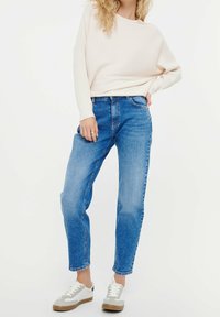 Kvinna iklädd en ljusbeige ribbad tröja, blå högmidjade jeans och vita sneakers, stående med ena handen på höften mot en vit bakgrund.