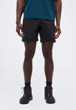 Mees, kellel on mustad sportlikud lühikesed püksid, mustad sokid ja mustad matkasaapad, seisab ühtlase heleda tausta ees.