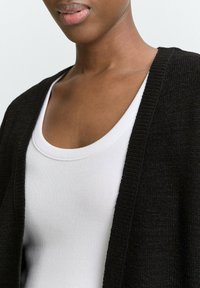 Schwarze Strickjacke mit offenem Vorderteil, kombiniert mit einem weißen, gerippten Tanktop. Texturiertes Material, lockere Passform und schlichte Gestaltung.