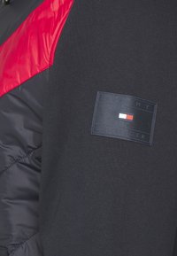 Giacca nera e rossa con texture trapuntata, polsini a coste e una toppa rettangolare con "TOMMY HILFIGER" e colori vivaci.