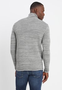 Grau gestrickter Pullover mit einem gerippten Muster, hohem Kragen und langen Ärmeln, getragen über blauen Jeans. Helle und dunkle Streifen bilden ein Muster.