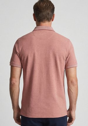 Man met kort haar, gekleed in een roze polo met korte mouwen en donkere broek, staand voor een effen lichtgrijze achtergrond.