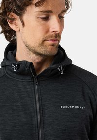 Zwarte zip-up hoodie gemaakt van gestructureerde stof met een strakke capuchon. Bevat een logo op de borst en trekkoorden op de capuchon.