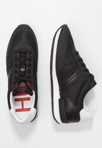 Chaussures de sport en maille noire avec des accents rouges, présentant un dessus texturé, une semelle intermédiaire blanche et un talon noir lisse. Lacées avec six œillets.