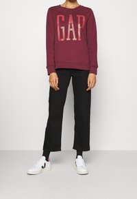 Sudadera burdeos con un gran logo "GAP", combinada con pantalones de pierna ancha negros y zapatillas blancas. Diseño simple y casual con puños acanalados.