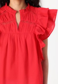Blouse en coton rouge avec manches à volants, encolure en V, et délicats détails de dentelle horizontale aux épaules. Taille froncée pour une coupe ample.