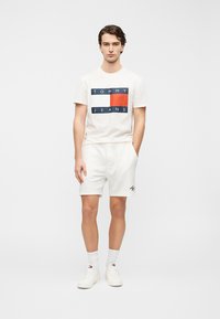 Λευκό βαμβακερό T-shirt με γαλάζιο και κόκκινο γραφικό "TOMMY JEANS", συνδυασμένο με λευκά σορτς με κορδόνι και λευκά αθλητικά παπούτσια με λογότυπα.