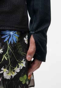 Svart og grønn langermet topp med perforert tekstur, kombinert med et blomsterprintet, mørkt stoffskjørt, som viser håndplassering ved sømmen.