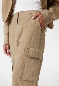 Beige cargo-housut ryppyisellä tekstuurilla, joissa on joustava vyötärö, sivutaskut ja vetoketjullinen tasku säilytystä varten.