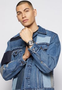 Veste en denim bleu avec un design patchwork, présentant différentes nuances de bleu et une montre noire avec un cadran rond et un bracelet noir.