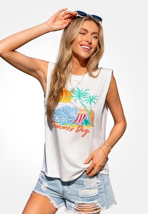 Femme souriante portant un débardeur blanc sans manches "Summer Day" avec un imprimé tropical, un short en jean déchiré et ajustant des lunettes de soleil sur sa tête.