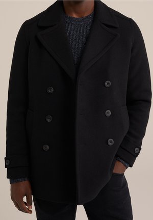 Mężczyzna ubrany w czarny dwurzędowy płaszcz typu pea coat z guzikami, w zestawie z ciemnym, teksturowanym swetrem i czarnymi spodniami.