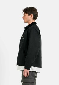 Giacca nera in tessuto con colletto, dotata di una tasca sul petto e tasche laterali con zip, indossata sopra una camicia grigia e pantaloni cargo grigi.