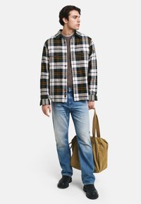 Koszula flanelowa w kratę w kolorze zielonym, białym i pomarańczowym, noszona na szarym topie, w połączeniu z jasnoniebieskimi dżinsami i czarnymi butami, trzymająca beżową torbę.